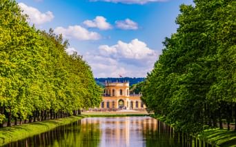 Yellow Orangerie palace in Kassel framed by symmetrical rows of green trees along a reflective canal under a blue sky with white clouds.