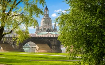 View of Dresden's Frauenkirche dome behind Augustus Bridge over the Elbe River, framed by green trees and lawn on a sunny day.