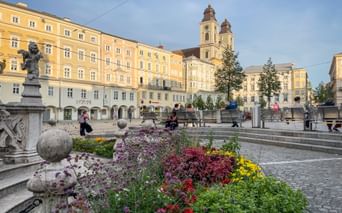 Linzer Hauptplatz mit bunten Blumenbeeten im Vordergrund, gelben historischen Gebäuden und Kirche mit zwei Türmen unter blauem Himmel.