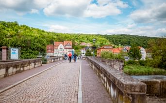 Two cyclists riding across historic cobblestone Werra bridge in Hann. Münden with colorful half-timbered houses and green hills in background.