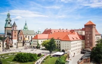 Aerial view of Wawel Castle complex in Krakow with red-roofed buildings, cathedral with green copper towers, and round brick tower.