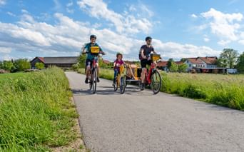 Family of four cycling on paved path through green countryside. Two adults and two children with trailer bike under blue sky with clouds.