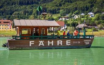 Fähre mit Radfahrern überquert die grüne Donau bei Engelhartszell. Bewaldete Hügel und Dorfgebäude im Hintergrund sichtbar.