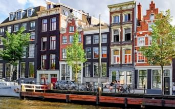 Row of traditional Amsterdam canal houses in various colors along a waterway. Bicycles are parked on a wooden dock in front of the buildings.