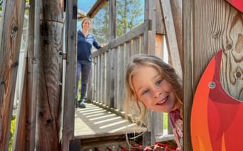 Smiling girl peeks through wooden playground structure while woman stands on elevated walkway in background. Sunny day with blue sky and trees.