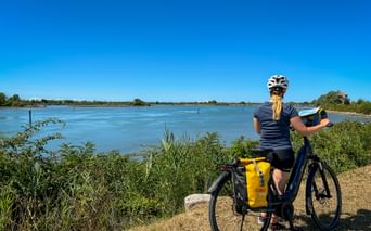 Radfahrerin mit Helm und gelben Packtaschen am Fahrrad blickt auf breiten Fluss unter blauem Himmel auf der Route Venedig-Mantua.