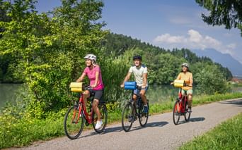 Three cyclists with helmets riding along a riverside path. Green trees line the route with mountains visible in the background under blue sky.