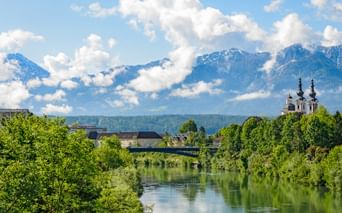 Blick auf die Drau in Villach mit einer Kirche mit zwei Türmen, grünen Bäumen und schneebedeckten Bergen unter blauem Himmel.
