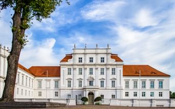 White baroque palace in Oranienburg with orange-tiled roof, symmetrical facade, and central entrance. A large tree stands in the foreground.