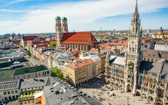 Aerial view of Munich's historic center showing Marienplatz with the New Town Hall tower and Frauenkirche's twin domes under blue sky.