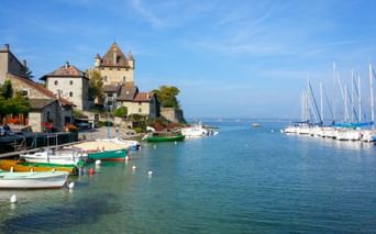 Malerischer Hafen von Yvoire am Genfersee mit bunten Booten, mittelalterlichen Schlosstürmen und Segelbooten unter blauem Himmel.