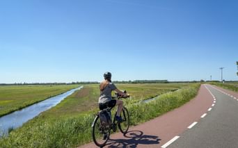 Radfahrerin auf rotem Radweg entlang Kanal durch flache grüne Polderlandschaft unter blauem Himmel zwischen Brügge und Amsterdam.