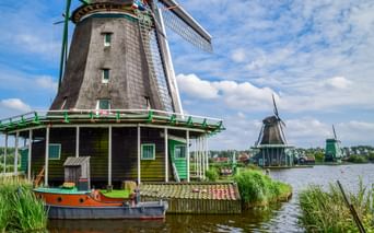 Traditionelle niederländische Windmühle mit grünem Rand neben einem Kanal in Kinderdijk. Ein kleines Boot liegt am Fuß vertäut, weitere Windmühlen sind in der Ferne sichtbar.