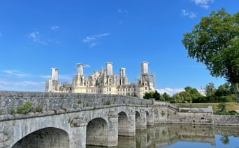 Stone bridge with multiple arches spanning calm water, leading to Château de Chambord with its distinctive towers under blue sky.