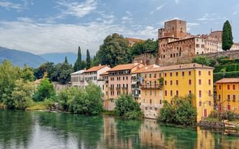 Farbenfrohe historische Gebäude von Bassano del Grappa spiegeln sich im Fluss Brenta, mit einer mittelalterlichen Burg auf dem Hügel.