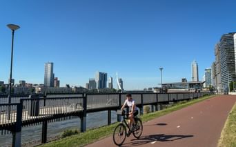 Radfahrerin auf rotem Radweg entlang der Nieuwe Maas in Rotterdam mit Skyline und Erasmusbrücke unter blauem Himmel.