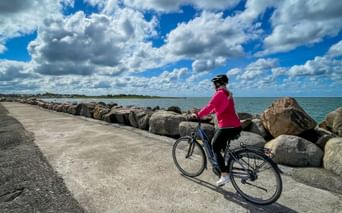 Radfahrerin in rosa Jacke mit Fahrrad auf gepflastertem Küstenweg neben steinerner Uferbefestigung. Blaues Meer und dramatischer Wolkenhimmel im Hintergrund.