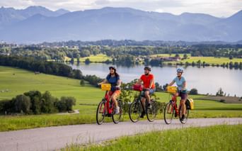 Three cyclists with yellow panniers on a path overlooking Riegsee lake, green meadows, and Bavarian Alps in the background under cloudy skies.
