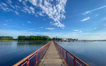 Long wooden pier with red railings extending across calm blue water near Cammin, with green shoreline and town buildings visible in the distance.