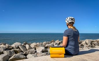 Radfahrerin mit weißem Helm sitzt auf der Promenade in Lido di Venezia mit Blick aufs Meer. Gelbe Eurobike-Taschen neben ihr, Steinmole sichtbar.