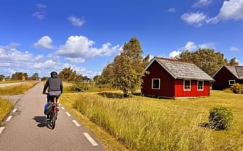 Radfahrer auf asphaltiertem Radweg entlang Sydkustleden in Schweden, vorbei an traditionellem roten Holzhaus mit weißen Rahmen unter blauem Himmel.