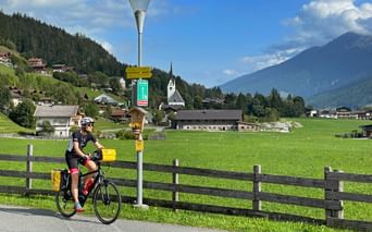 Radfahrer am Tauernradweg bei Krimml mit grünen Wiesen, Alpendorf mit Kirche, Holzzaun und Bergen unter blauem Himmel.