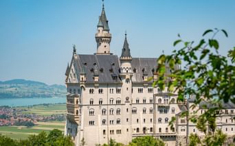 Schloss Neuschwanstein mit weißen Mauern und grauen Türmen umgeben von grünen Bäumen. See und Dorf im Hintergrund unter blauem Himmel.