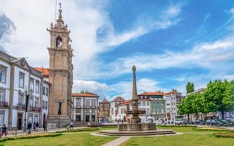 Largo Carlos Amarante in Braga with bell tower, fountain, green lawns, and colorful buildings under blue sky.