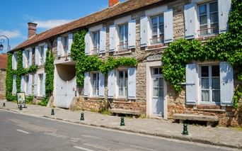 Two-story stone building in Barbizon with white shutters and ivy-covered walls. The facade features multiple windows and a central entrance.