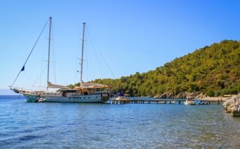 Weißes Segelschiff MS Boreas am Holzsteg in ruhiger Bucht. Bewaldeter Hang und klares blaues Wasser im Golf von Gökova, Türkei.
