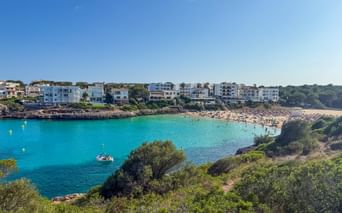 Turquoise bay of Cala Marcal beach in Porto Colom, Mallorca, with white buildings along the shore and green vegetation in foreground.