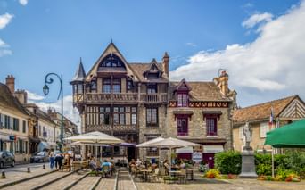 Half-timbered Maison Raccollet building in Moret-sur-Loing with outdoor café seating on a cobblestone square under blue sky.