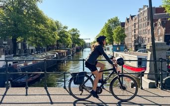 Radfahrerin auf Brücke mit Blick auf Amsterdamer Gracht mit Hausbooten, traditionellen niederländischen Gebäuden und Bäumen am Ufer.