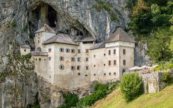 Burg Predjama in eine massive Kalksteinklippe in Slowenien gebaut, mit Höhleneingang darüber. Grüne Wiese im Vordergrund, Bäume rechts.