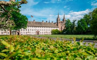 Schloss Corvey in Höxter mit Doppelturmkirche und Schlossflügel, umgeben von grünen Rasenflächen und Bäumen unter blauem Himmel.