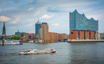 Hamburger Hafen mit Elbphilharmonie, Ausflugsbooten auf der Elbe und roten Backsteingebäuden unter bewölktem Himmel.