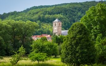 Historic tower of Creuzburg Castle rising above red-roofed buildings, surrounded by lush green trees and forested hills in Thuringia.