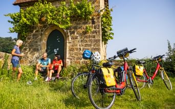 Drei Radfahrer rasten im Schatten einer Steinkapelle umgeben von Weinreben. Drei Fahrräder mit gelben Taschen stehen im Gras.