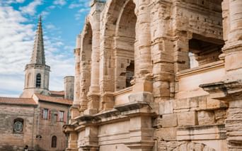 Ancient Roman amphitheater in Arles with weathered stone arches and columns. A church tower with red-tiled roofs visible in the background.