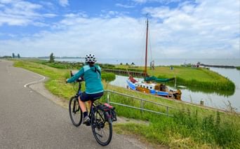 Cyclist in turquoise jacket riding on dike path alongside canal with moored sailboat. Green meadows and blue sky with clouds visible.