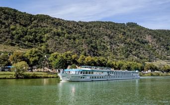 MS Switzerland Flusskreuzfahrtschiff fährt auf ruhigem grünen Wasser mit bewaldeten Hügeln im Hintergrund unter teilweise bewölktem Himmel.