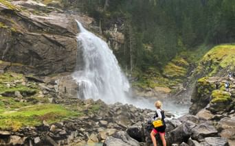 Cyclist with yellow backpack viewing Krimml Waterfalls cascading over rocky cliffs surrounded by green forest along the Tauern cycle path.