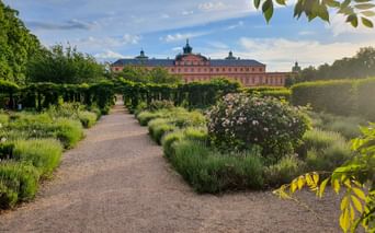 Kiesweg durch barocken Garten mit Lavendelbeeten und Rosenbüschen führt zum roten Schloss Rastatt unter blauem Himmel.