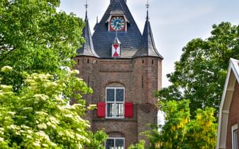 Historisches Stadttor Lekpoort in Vianen mit roten Backsteinmauern, Schieferdach, Uhr und roten Fensterläden, eingerahmt von grünen Bäumen.