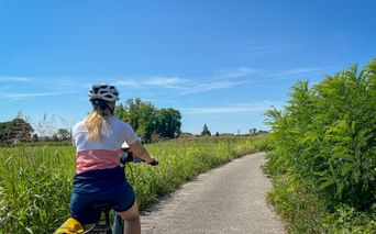 Radfahrerin mit blondem Pferdeschwanz und Helm auf asphaltiertem Weg durch grüne Wiesen mit hohen Gräsern und Farnen unter blauem Himmel.