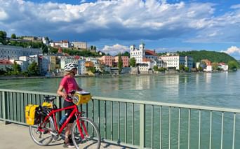 Radfahrerin mit rotem Fahrrad und gelben Packtaschen auf Brücke mit Blick auf Passauer Stadtbild entlang der Donau unter blauem Himmel.