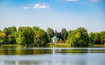 View across a calm pond reflecting a white pavilion with turquoise dome surrounded by lush green trees under blue sky in Kassel.