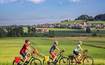 Three cyclists with panniers riding through green countryside near Michaelbeuern. Village with church visible on hillside in background.