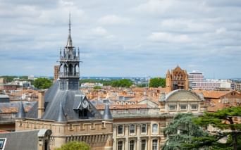 Aerial view of Toulouse with the Donjon du Capitole tower prominently featured. Red-tiled rooftops and historic buildings under a cloudy sky.
