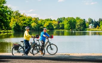 Two cyclists with e-bikes pause by a lake in Karlsaue Park, Kassel. A white pavilion is visible across the water surrounded by green trees.